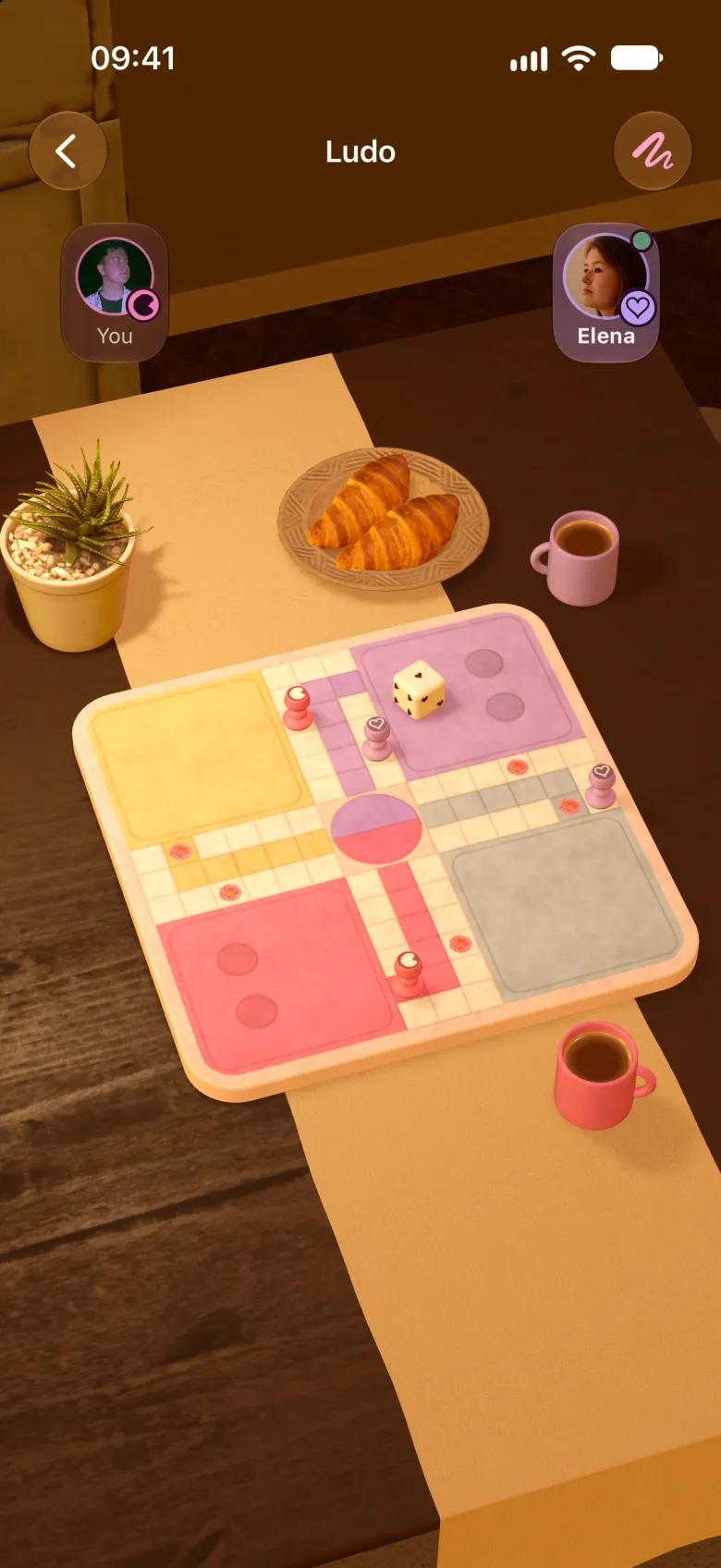 A game of Ludo on a wooden table with croissants, a small plant and a mug of hot chocolate for each partner. Each partner's pieces carry the mark they drew.